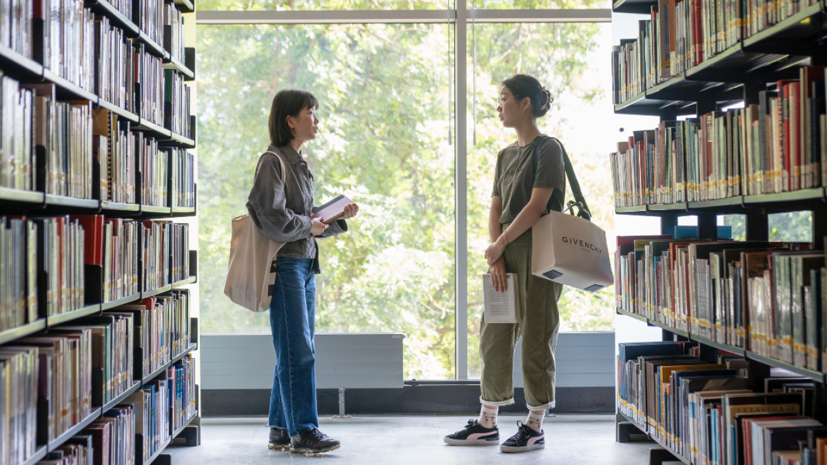 Two students in the library, standing next to bookshelves