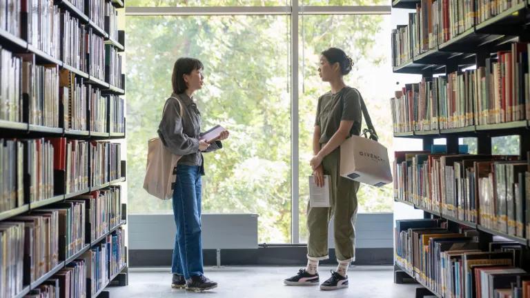 Two students in the library, standing next to bookshelves